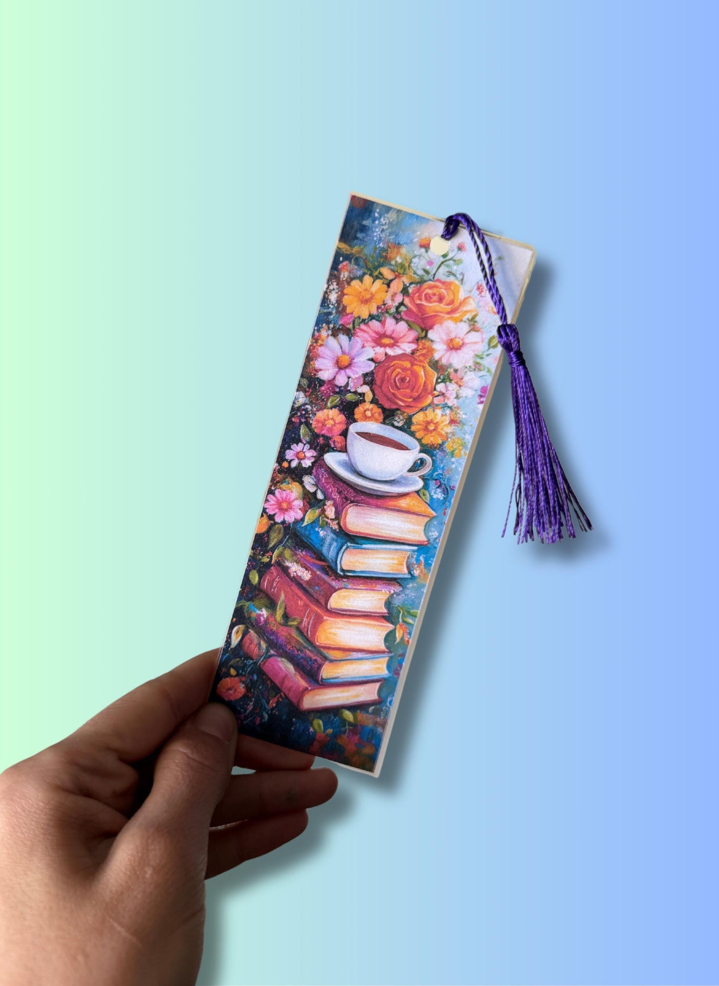 A hand holds up the laminated floral bookmark against a soft green and blue gradient background, showing the glossy finish, the book stack and teacup design, and the purple tassel. __etsy:6777949668