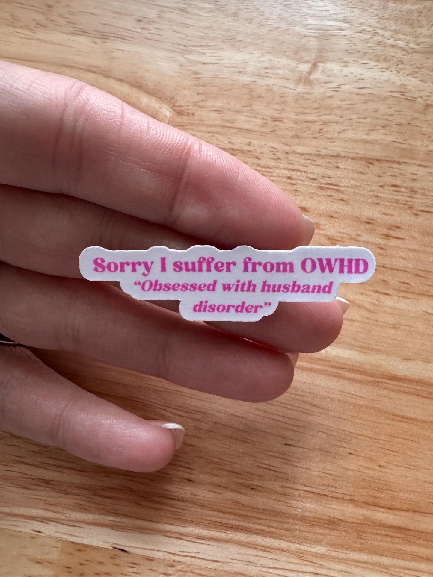 A hand holds the physical "Sorry I suffer from OWHD - Obsessed with husband disorder" die-cut sticker between two fingers over a light wood surface, showing its true size and glossy pink-and-purple finish. __etsy:6706928777