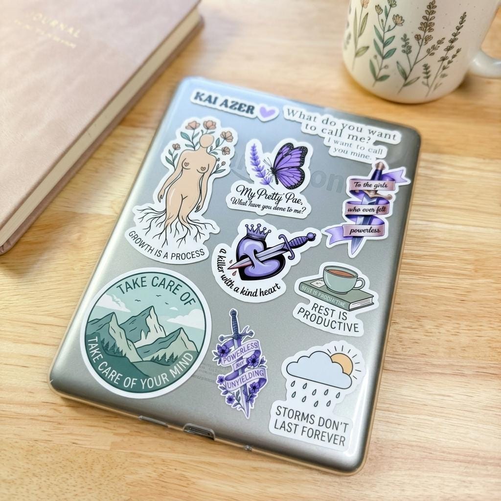 The back of a silver laptop densely covered in book boyfriend vinyl stickers, on a wooden desk with a floral journal and a botanical mug. Mental-health themed decals include Take Care of Your Mind with mountains, Rest is Productive, Storms Dont Last Forever, Growth is a Process, a butterfly, a crowned heart with dagger, and the name Kai Azer. __etsy:8133863942