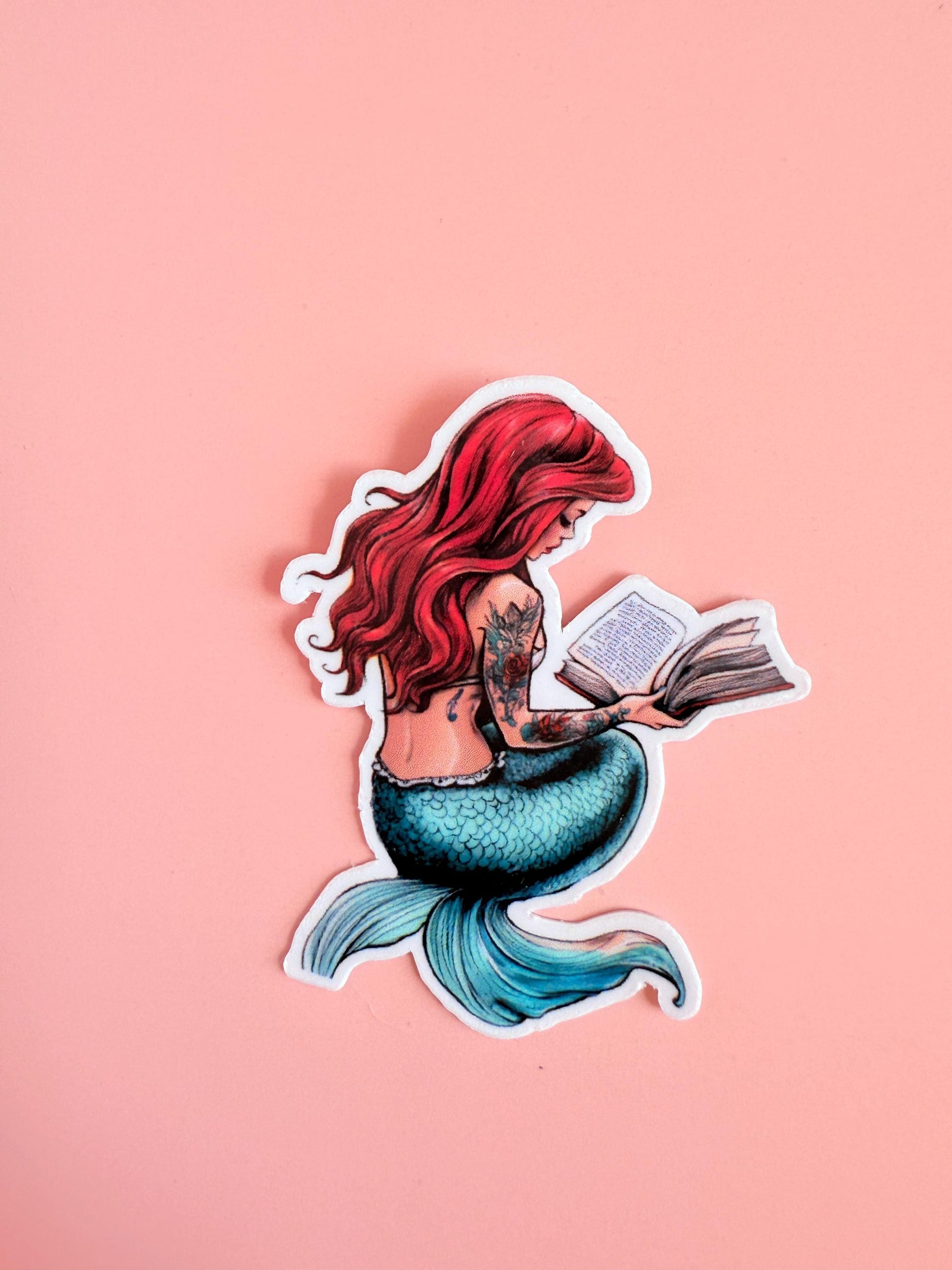 Red-haired mermaid with teal tail, sitting and reading a book. Tattooed arm visible. Vibrant fantasy sticker on a soft pink background. __etsy:6940969969