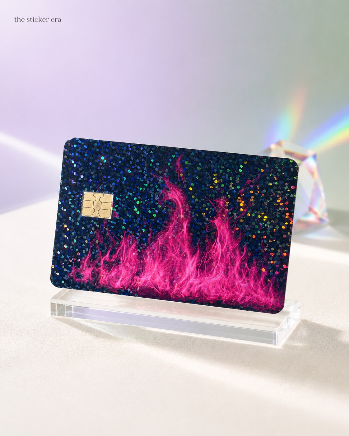 Holographic glitter pink fire credit card skin catching rainbow light on a clear stand by The Sticker Era
