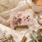 A rectangular pink glitter pouch with a debit card skin print and a small gold tag, displayed on a gold stand on a cream fabric surface with an open book, dried flowers, a cup of coffee, and greenery. by the sticker era