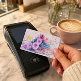 Hand holding a pastel floral book card skin by a black payment terminal with a latte and a pink peony by The Sticker Era