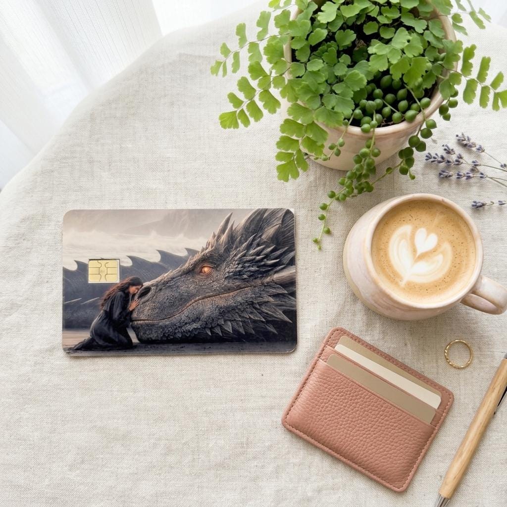 a debit card skin of a black dragon laying it's head down so a human can rest her head on the dragon on a cream colored cloth, a small green plant and a pinkish colored wallet on the side. 
and a hot coffee
 made by thestickerera __etsy:8156746631