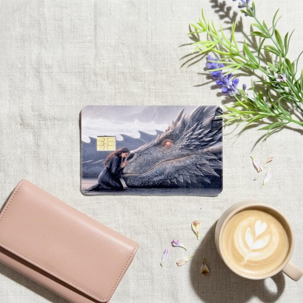 a debit card skin of a black dragon laying it's head down so a human can rest her head on the dragon on a cream colored cloth, a lavander 
plant and a pinkish colored wallet on the side. 
and a hot coffee
 made by thestickerera __etsy:8156746637