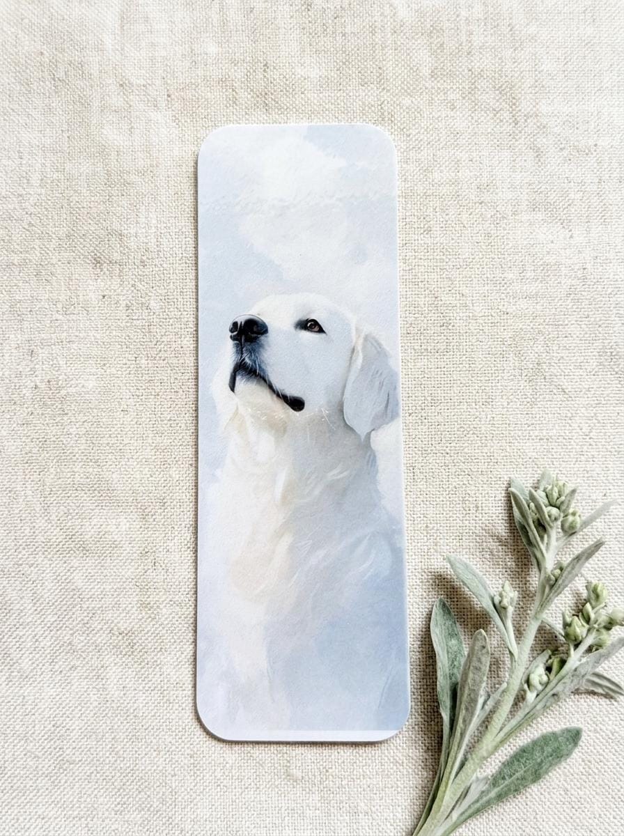 Close-up of a single English Cream Golden Retriever watercolor bookmark on a beige linen background, with a sprig of soft green foliage beside it. __etsy:8181674243