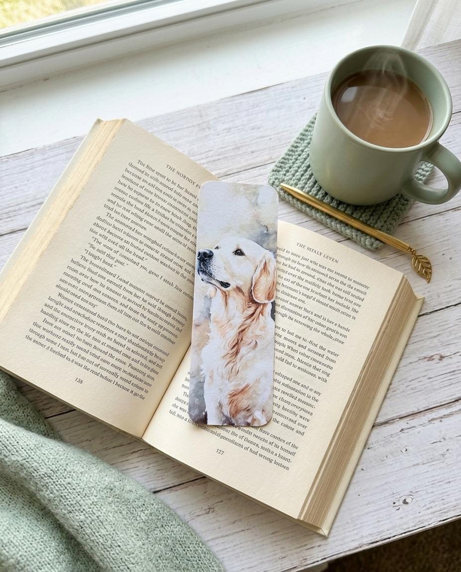 A single English Cream Golden Retriever watercolor bookmark resting on an open book, with a cup of tea, a gold pen, and a knit blanket on a wooden surface near a window. __etsy:8133771646