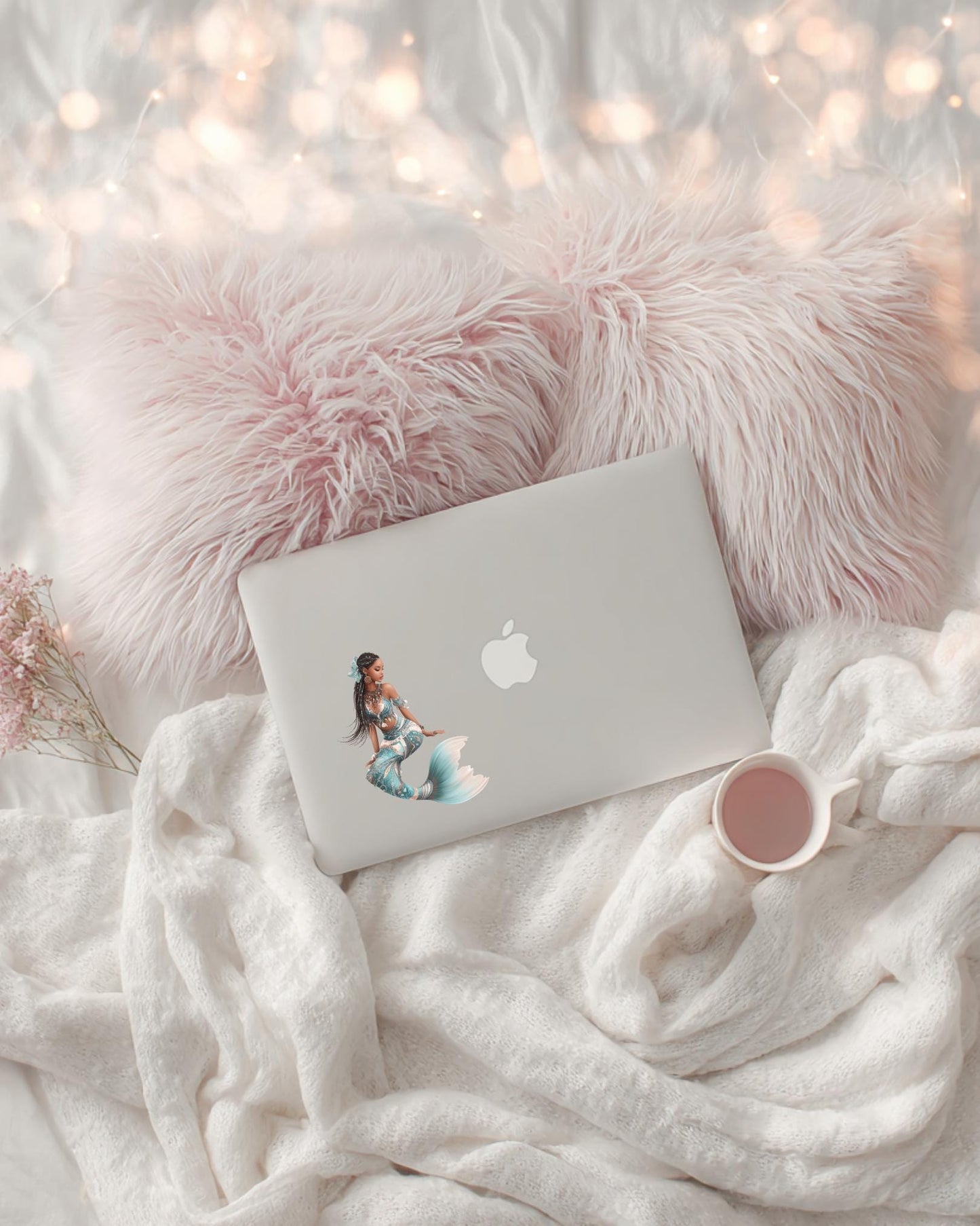 A white laptop decorated with the Ethiopian mermaid sticker, resting on a cozy cream blanket with a pink fur pillow, fairy lights and a pink mug. __etsy:7677847986