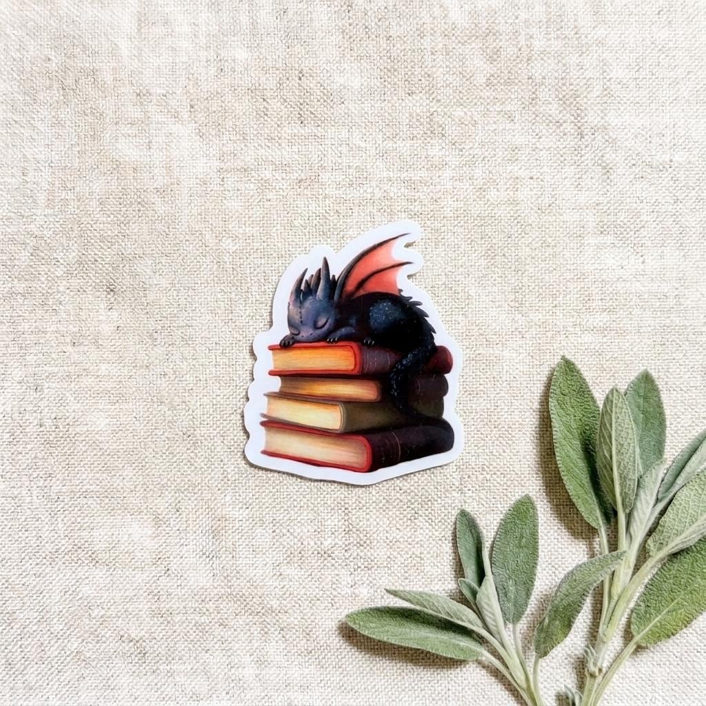 Book dragon vinyl sticker of a sleeping black baby dragon on a stack of colorful books, displayed on natural linen with fresh sage leaves. __etsy:8134203582