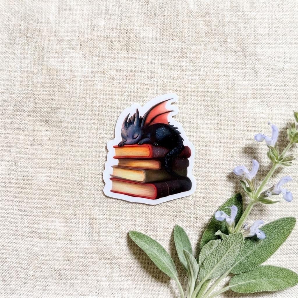 Die-cut vinyl sticker of a sleeping black baby dragon curled on a stack of colorful fantasy books, resting on a linen background beside fresh sage leaves and lavender blooms. __etsy:8134203586