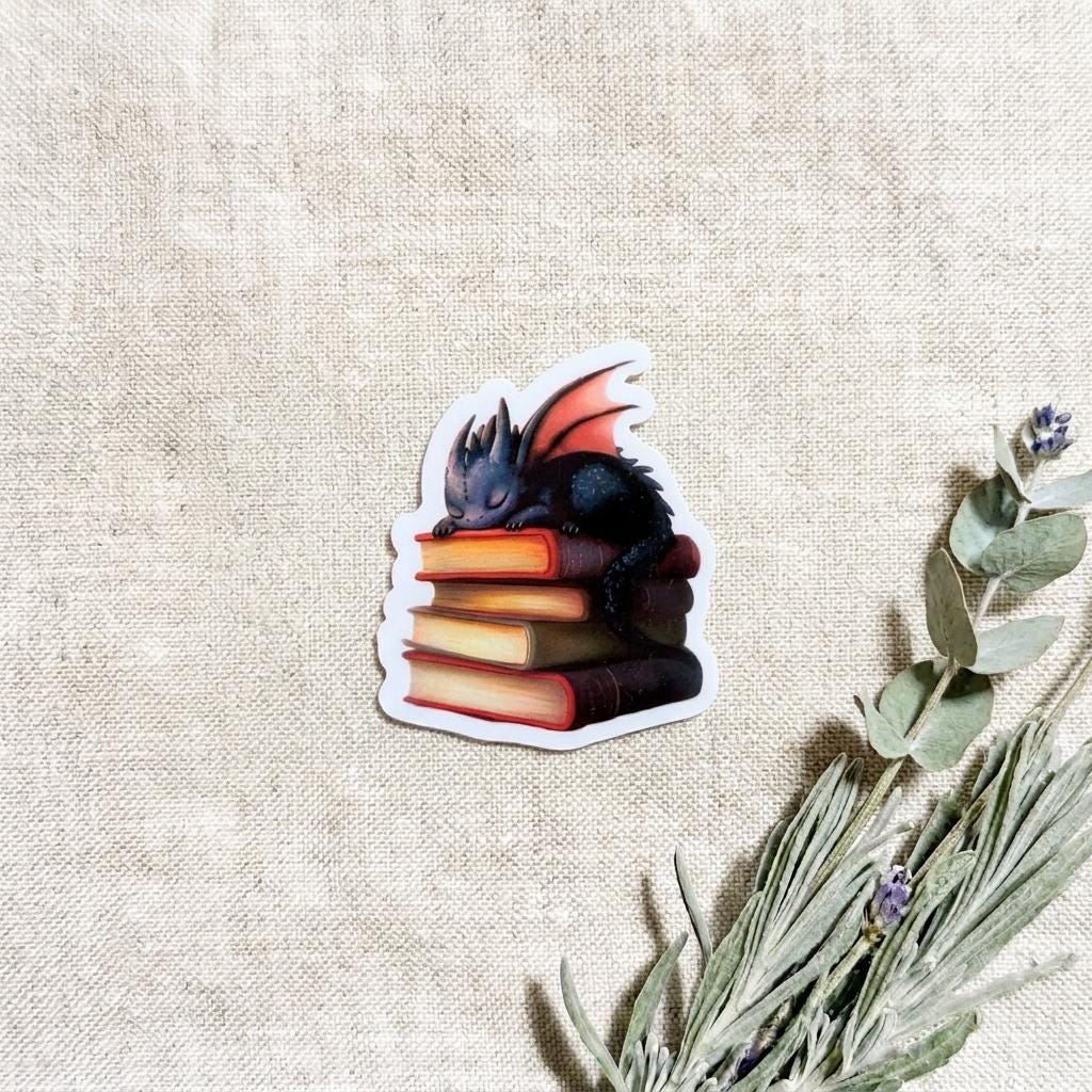 Close-up of the black baby dragon vinyl sticker sleeping atop a stack of colorful books, styled on natural linen with eucalyptus and lavender sprigs. __etsy:8182098539