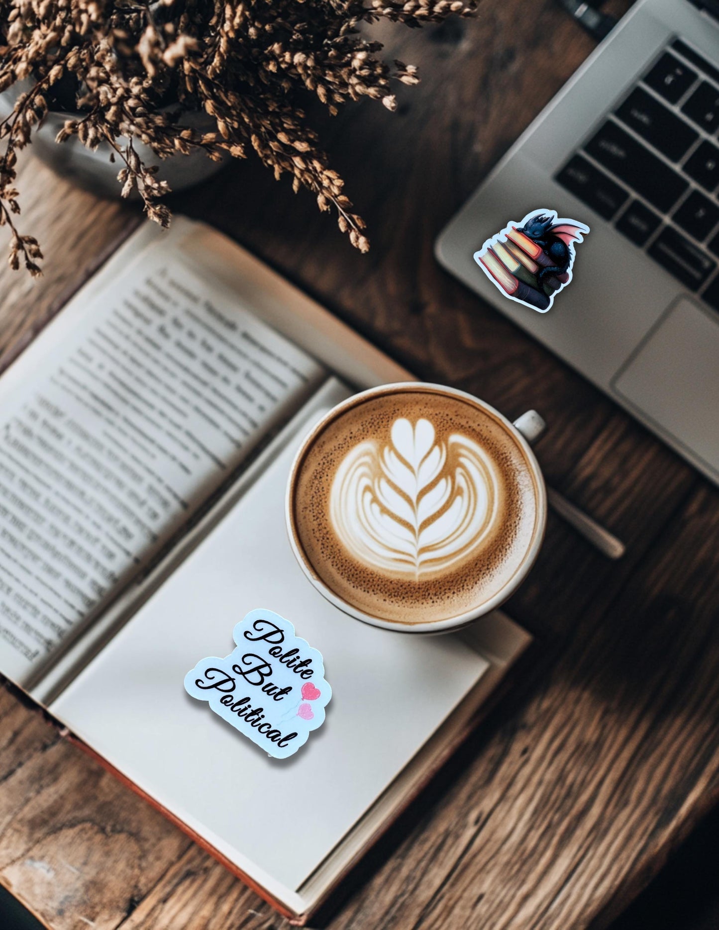 May include: A latte with latte art in a white mug on a book with a sticker that says "Polite But Political" with two pink Cozy reading flat-lay with an open book, a latte with leaf art, dried flowers, and a laptop featuring the book dragon sticker on a dark wood table.hearts. The book is on a wooden table with a laptop and a plant in the background. __etsy:6823101623