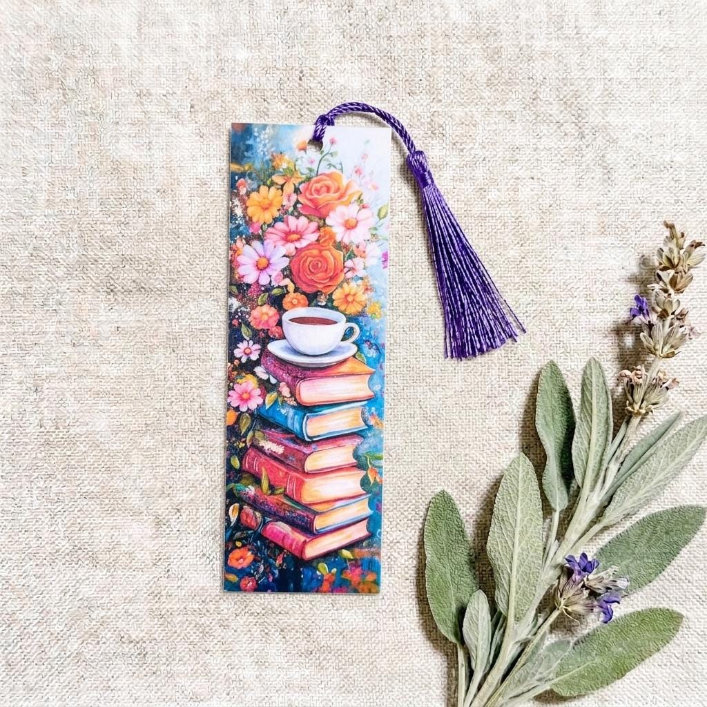Floral bookmark with a watercolor design of stacked books topped with a teacup and a cluster of roses and daisies, with a long purple tassel, shown on woven linen beside sage and lavender. __etsy:8182193831