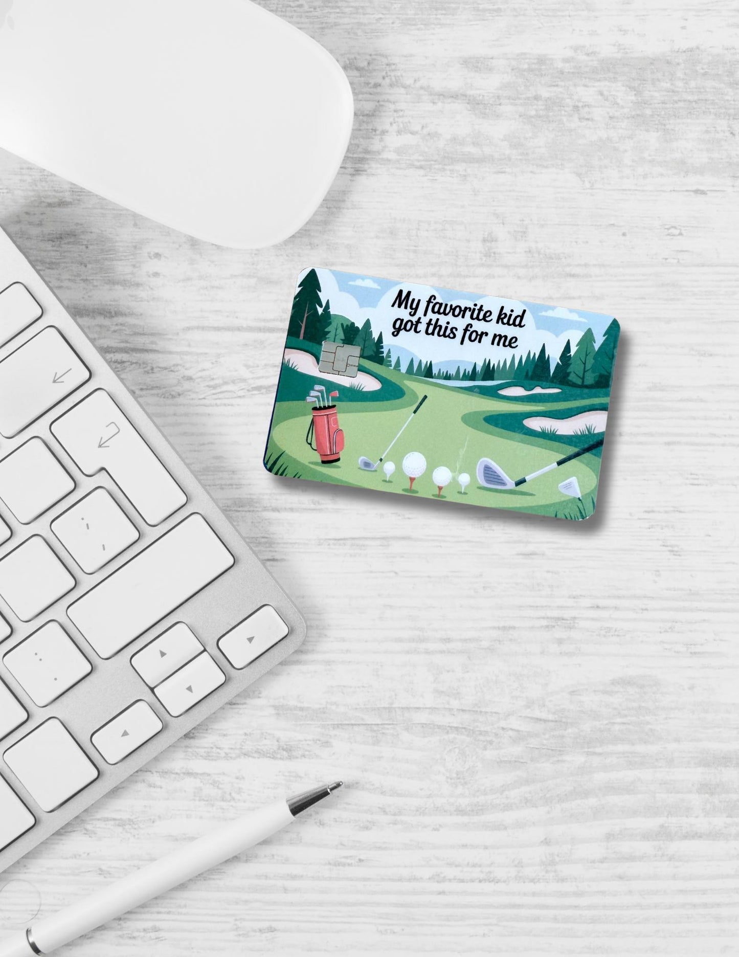A golf-themed debit card skin sits on a white wood desk next to a wireless mouse, computer keyboard, and pen. The card design features a green golf course, golf clubs, balls, and the humorous phrase "My favorite kid got this for me." It's a playful, sporty accessory with a lighthearted gift message perfect for Father's Day. __etsy:6864947806