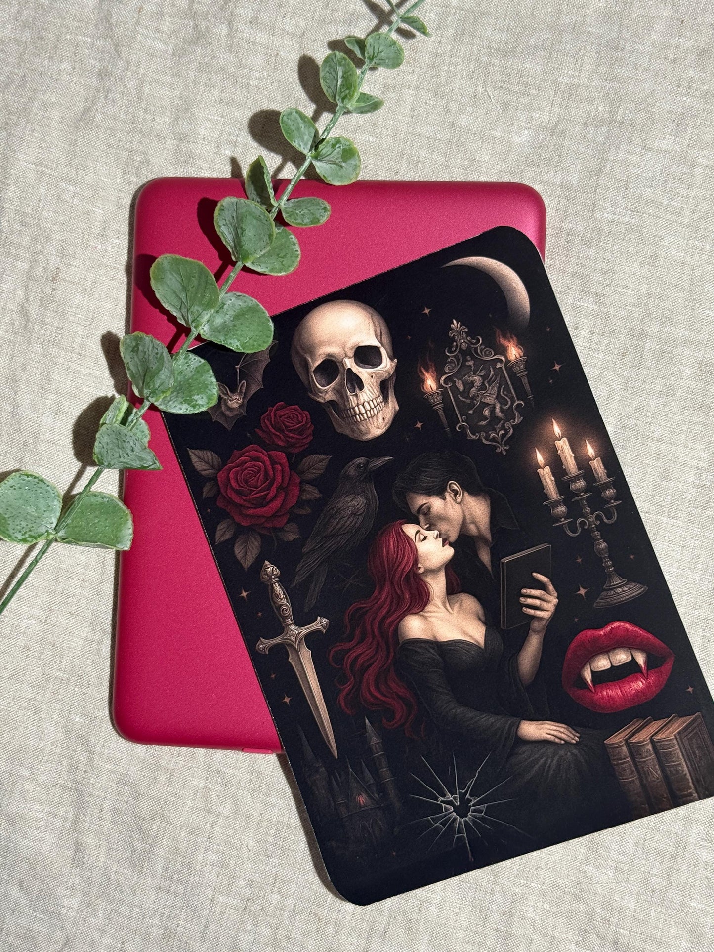 Gothic vampire Kindle insert decal applied to a pink e-reader, shown on a linen background with eucalyptus greenery. The dark romantasy design features a red-haired woman embracing a vampire, a skull, red roses, a raven, a dagger, and lit candelabra. __etsy:8165474921