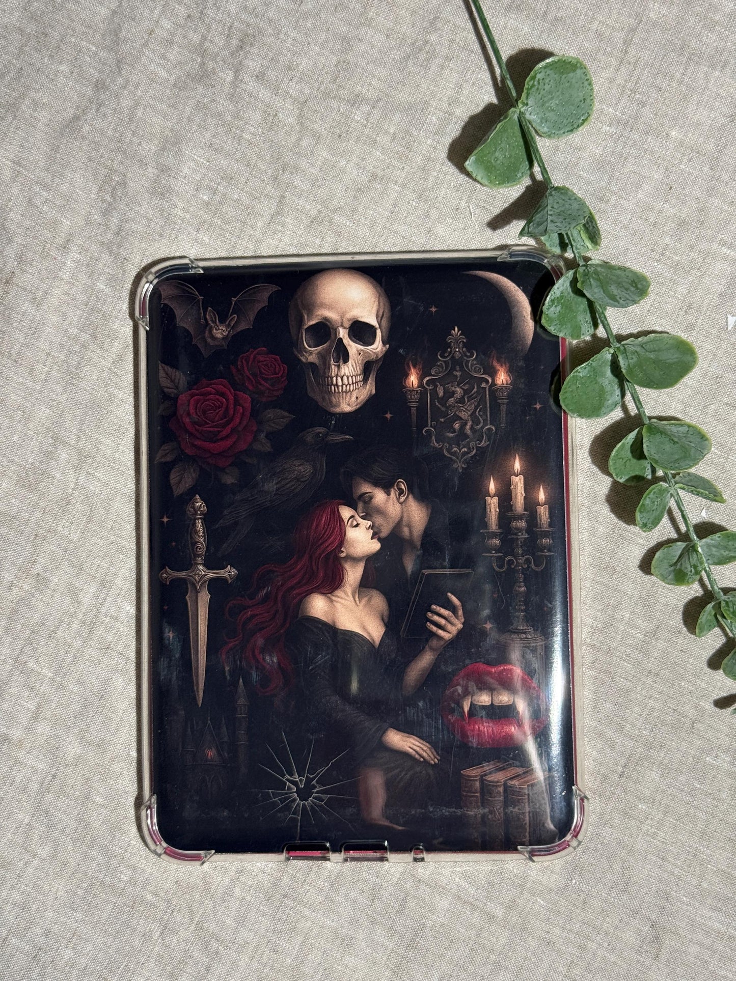 Gothic vampire Kindle insert decal displayed on a clear e-reader case with a trailing eucalyptus vine on a linen background. The dark romantasy illustration features a red-haired woman and a vampire embracing, a skull, red roses, a dagger, lit candelabra, and stacked books. __etsy:8117565746