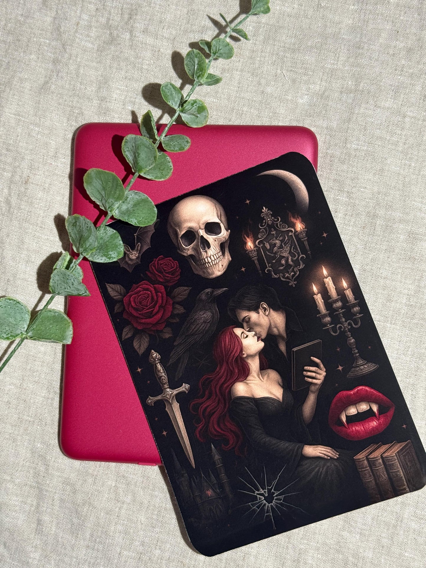 Gothic vampire Kindle insert decal angled over a pink e-reader with a trailing eucalyptus vine on linen. The dark romantasy illustration features a red-haired woman embracing a vampire, with a skull, red roses, a raven, a dagger, lit candelabra, and stacked books. __etsy:8117565732
