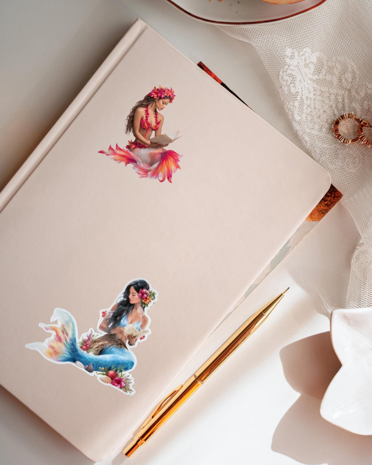 Two Hawaiian mermaid stickers, one with a red tail and one with a teal tail, placed on a cream notebook beside a gold pen in a flat lay __etsy:7732326287