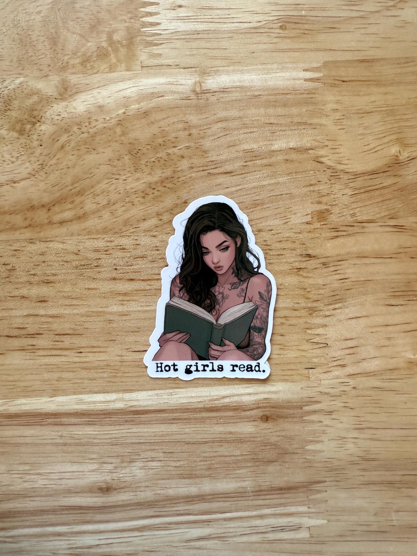 A sticker of a confident woman with dark hair and bold tattoos reading a book. A feminist and empowering design for readers. The sticker reads "hot girls read" __etsy:6708682352