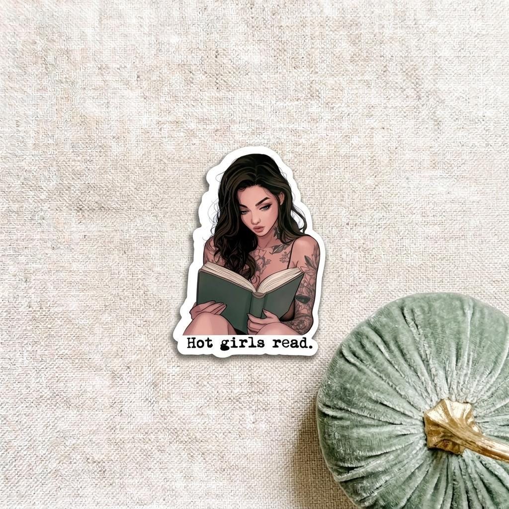 Die-cut vinyl sticker of an illustrated woman with long dark wavy hair and arm tattoos reading a green book, with the typewriter-style text "Hot girls read." beneath her. Shown on a linen background beside a sage green velvet pumpkin. __etsy:8134248698