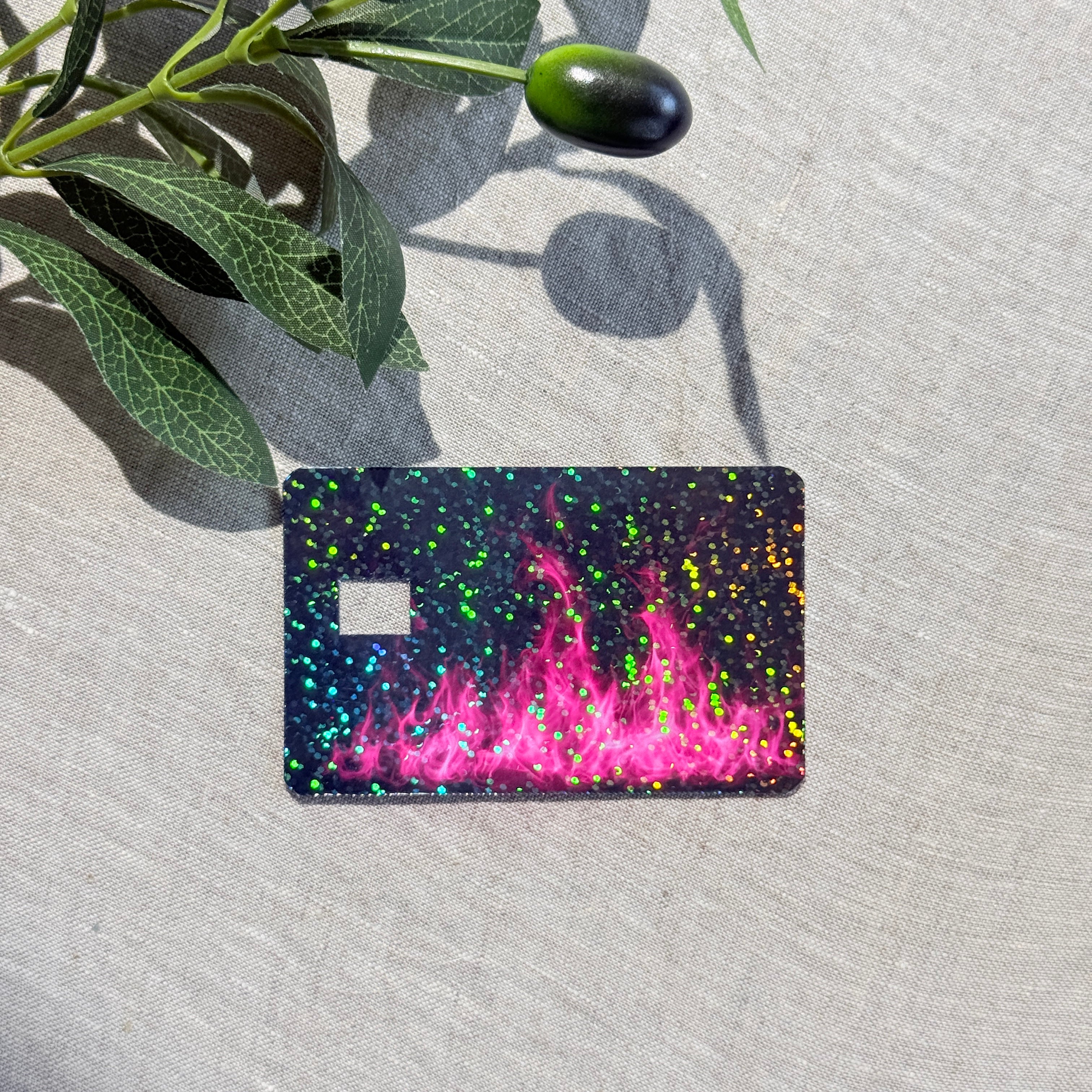 Hot pink flame glitter debit card skin lying flat under olive branch shadows by The Sticker Era