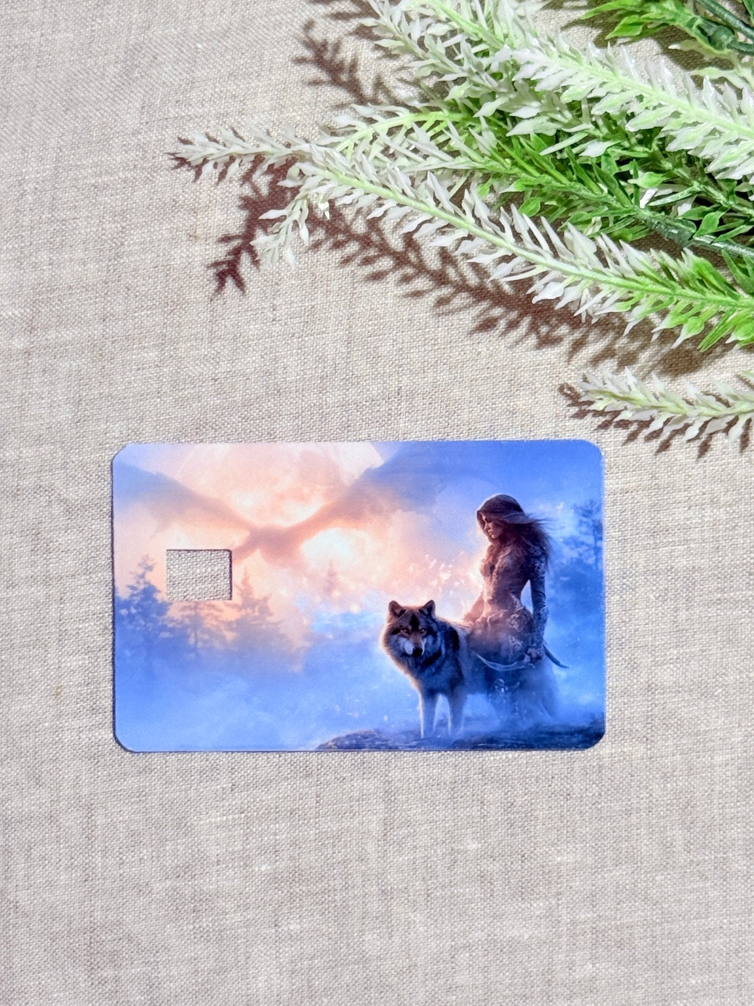Close-up of the transit-style wolf card with a rectangular swipe cutout, printed with the huntress and gray canine against a snowy blue and orange scene, lying on natural beige linen with a fern branch by The Sticker Era