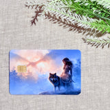 Full front view of the waterproof wolf debit card with gold chip lying flat on woven beige linen beside a green fern frond, showing the huntress, canine, snowy woodland, and glowing orange horizon by The Sticker Era