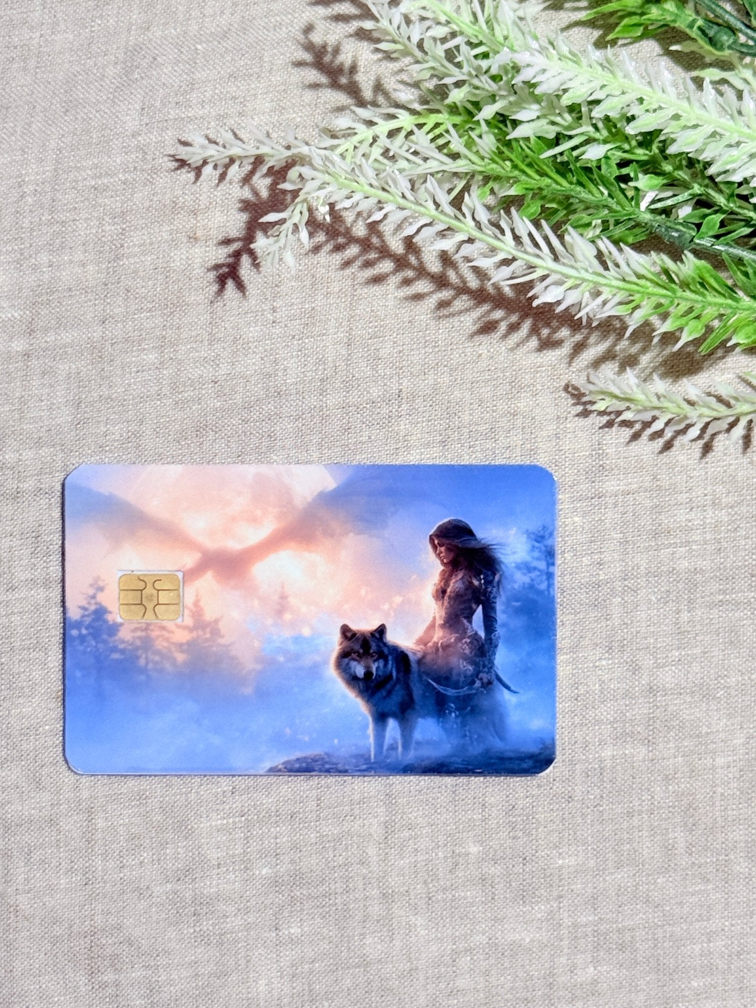 Full front view of the waterproof wolf debit card with gold chip lying flat on woven beige linen beside a green fern frond, showing the huntress, canine, snowy woodland, and glowing orange horizon by The Sticker Era