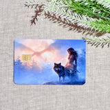 Angled product shot of the standard wolf card resting on textured oatmeal fabric with an evergreen sprig above, depicting the cloaked heroine and her companion beast on a rocky ledge beneath a fiery peach sunburst by The Sticker Era