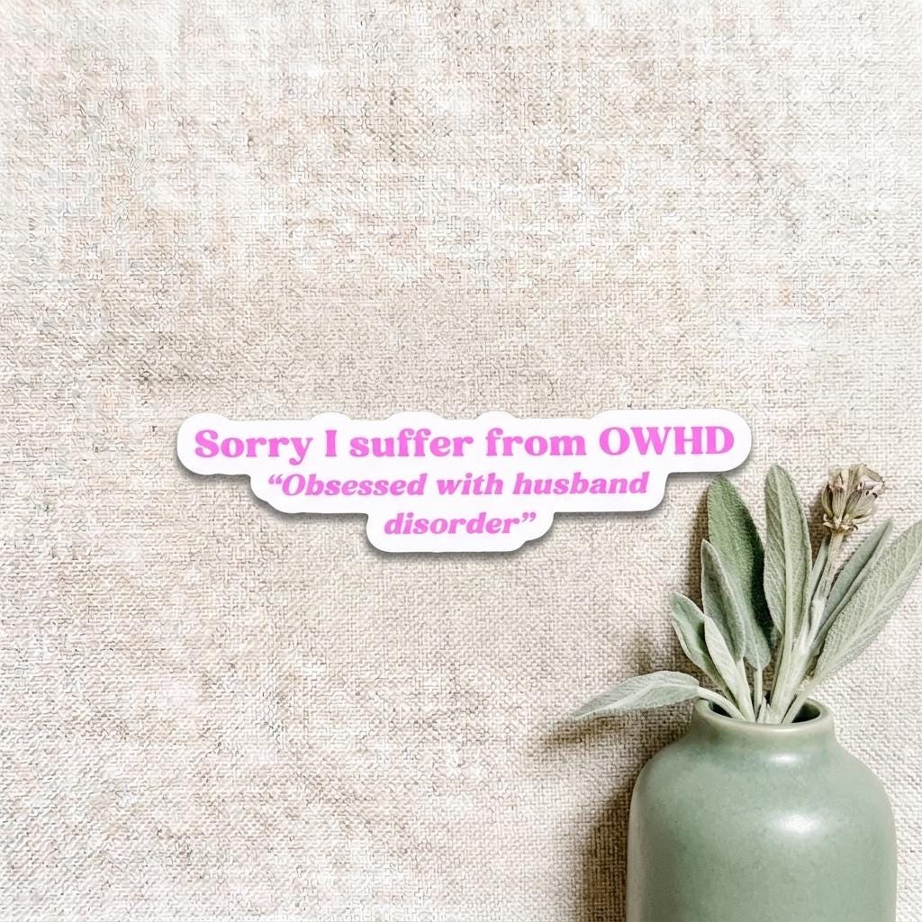 Pink and purple die-cut vinyl sticker reading "Sorry I suffer from OWHD - Obsessed with husband disorder" in cursive and bold lettering. Shown on a natural linen background beside a small sage green ceramic vase holding greenery. __etsy:8182190161