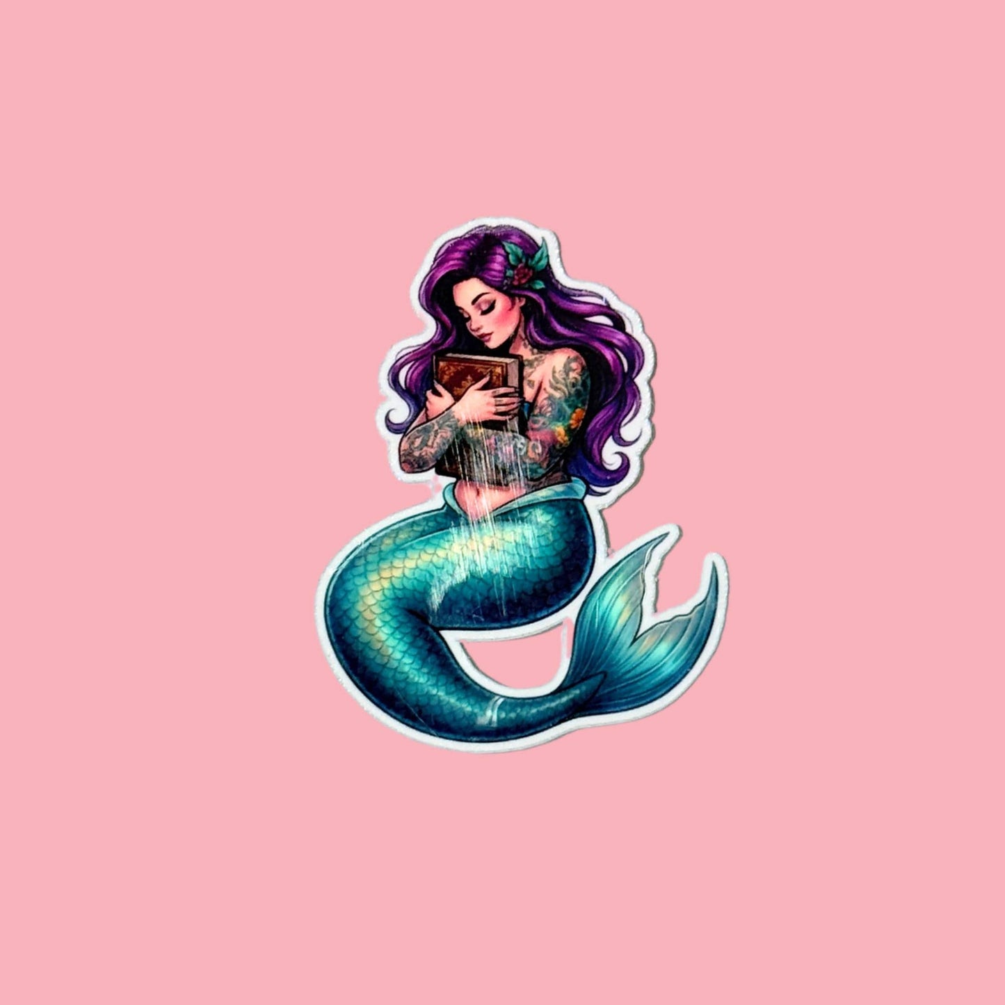 A single die-cut mermaid sticker on a solid pink background. The mermaid has long purple hair, tattooed arms, and a teal tail, and is shown holding a book. __etsy:7990868640