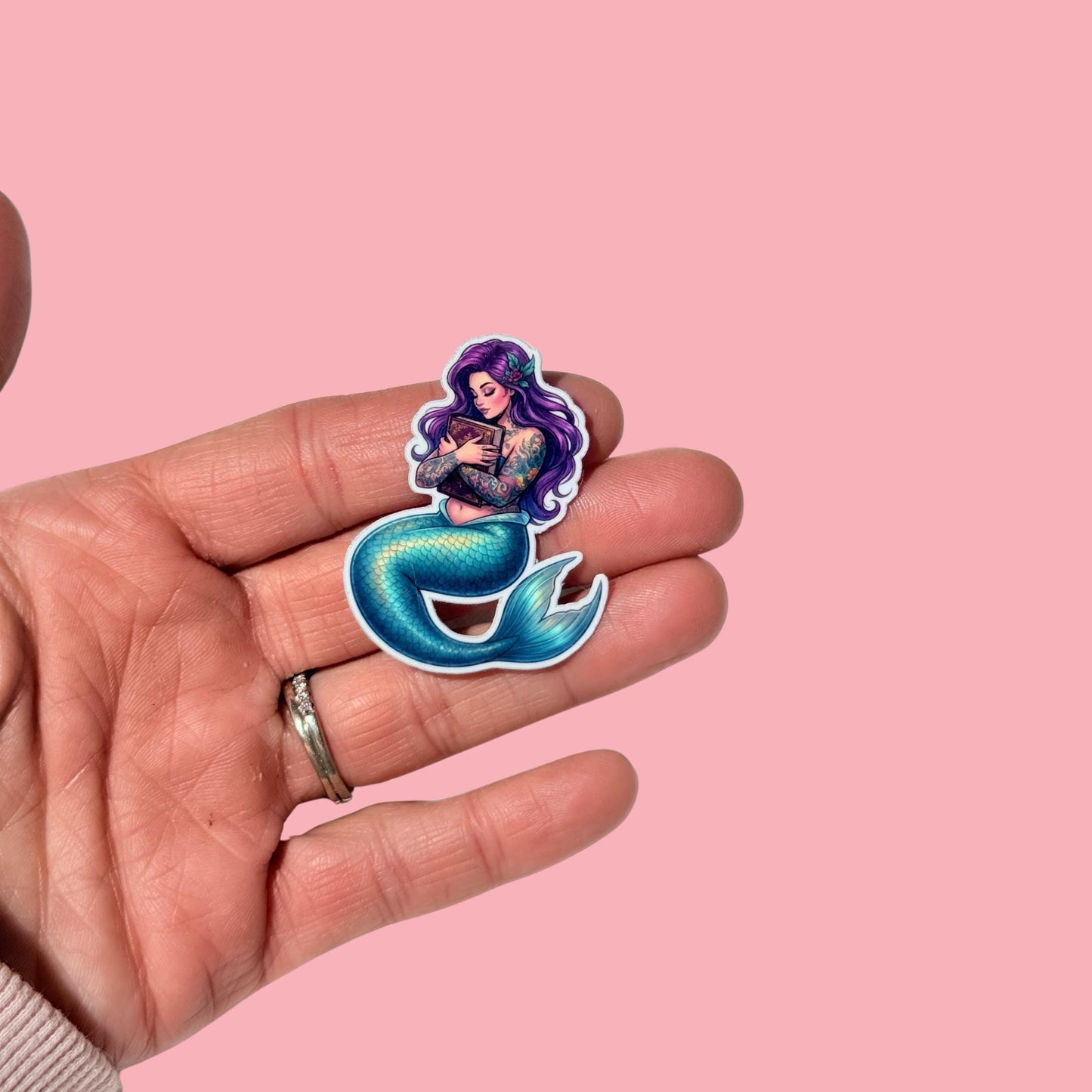 A hand holds a single mermaid sticker against a pink background, showing its size relative to the fingers. The mermaid has purple hair, tattooed arms, a teal tail, and is holding a book. __etsy:8038808143