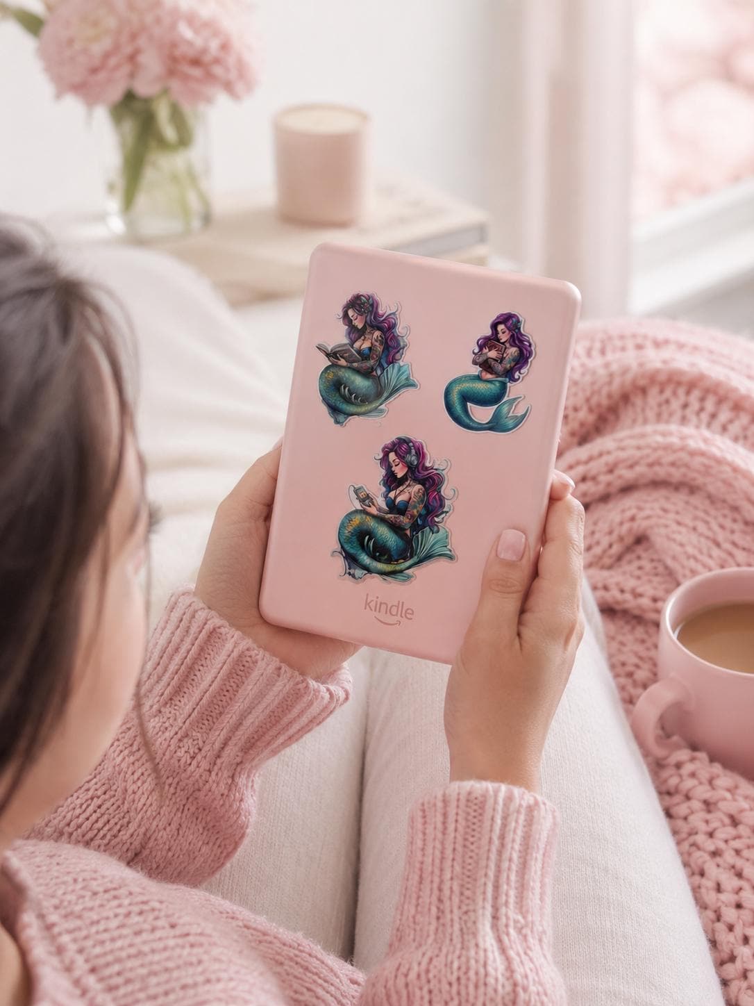 A person in a pink knit sweater holds a pink e-reader decorated with three purple-haired mermaid stickers. A cup of coffee and a vase of pink flowers sit nearby in a cozy setting. __etsy:7990888776
