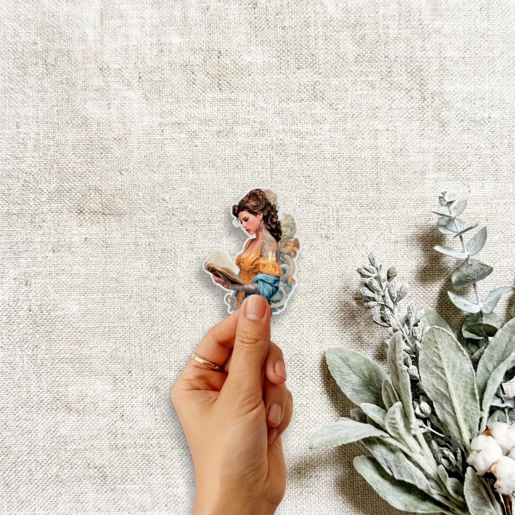 Hand holding a die-cut vinyl sticker of a brown-haired woman reading a book in a yellow and blue dress, with eucalyptus, lamb's ear, and cotton stems on a natural linen background. __etsy:8120643526