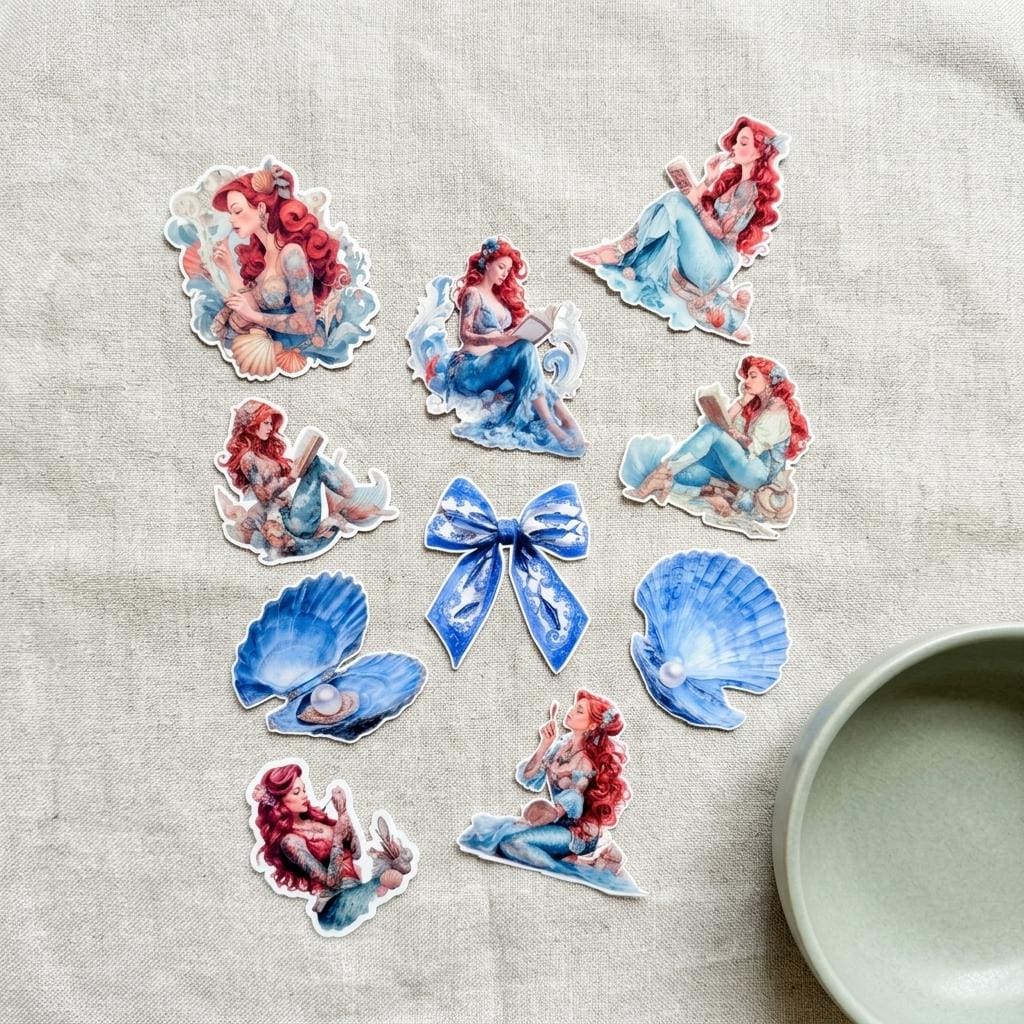 Full waterproof vinyl sticker pack laid flat on natural linen fabric, featuring tattooed mermaid decals with red hair and blue tails, a blue patterned bow sticker, and two blue seashell stickers, with a sage green dish in the corner. __etsy:8165922899