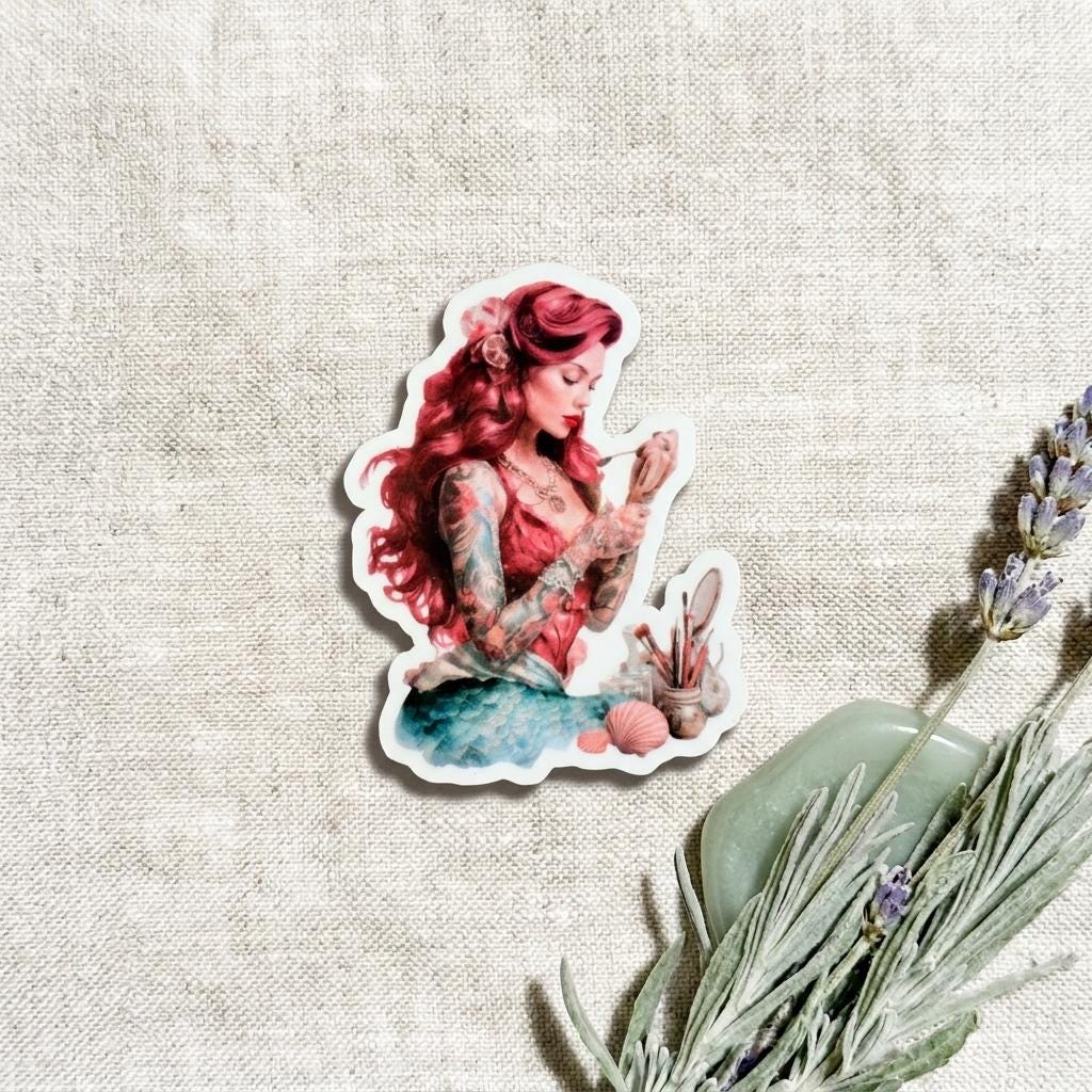 Close-up of a single waterproof vinyl sticker showing a red-haired tattooed mermaid seated with a blue-green tail and detailed arm and body tattoos, placed on linen beside dried lavender. __etsy:8118012820