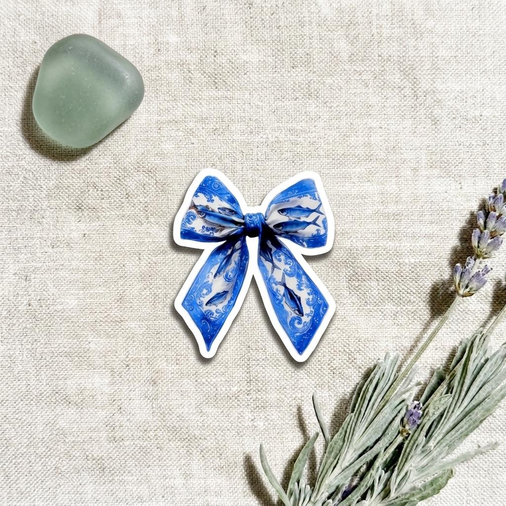 Close-up of a single waterproof vinyl sticker shaped like a blue and white patterned ribbon bow, styled on linen with sea glass and dried lavender nearby. __etsy:8165922897