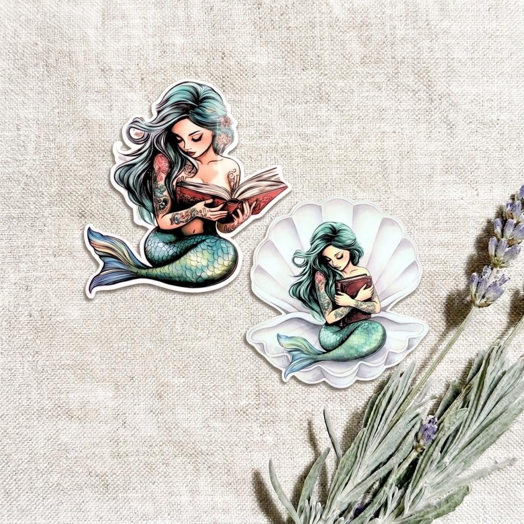 Two teal mermaid vinyl stickers on a linen background with lavender sprigs. One mermaid with flowing silver hair reads an open book; the other sits inside a white seashell holding a book, both with green-blue tails. __etsy:8181740589