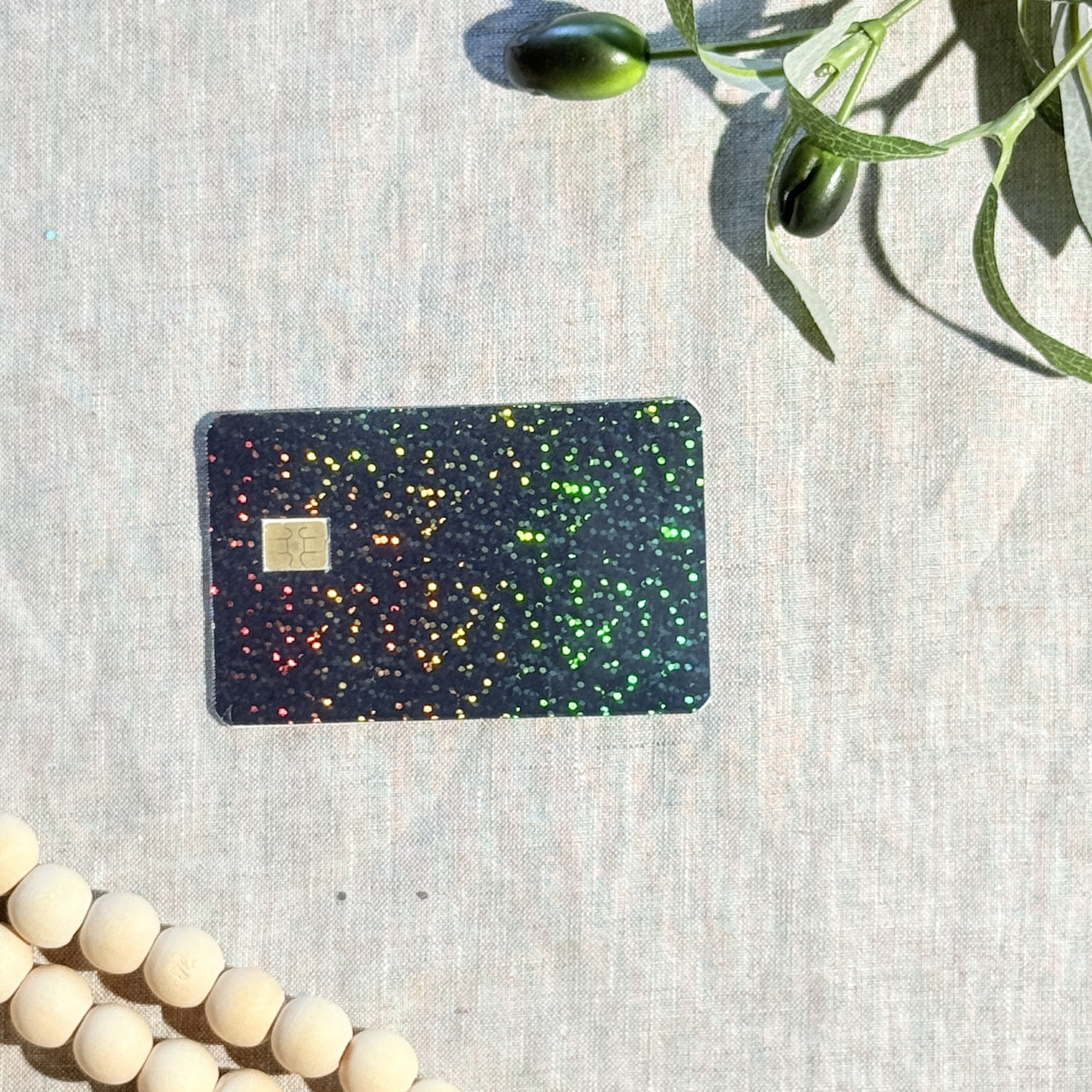 Black sparkle debit card skin catching rainbow light, flat lay on cream linen by The Sticker Era