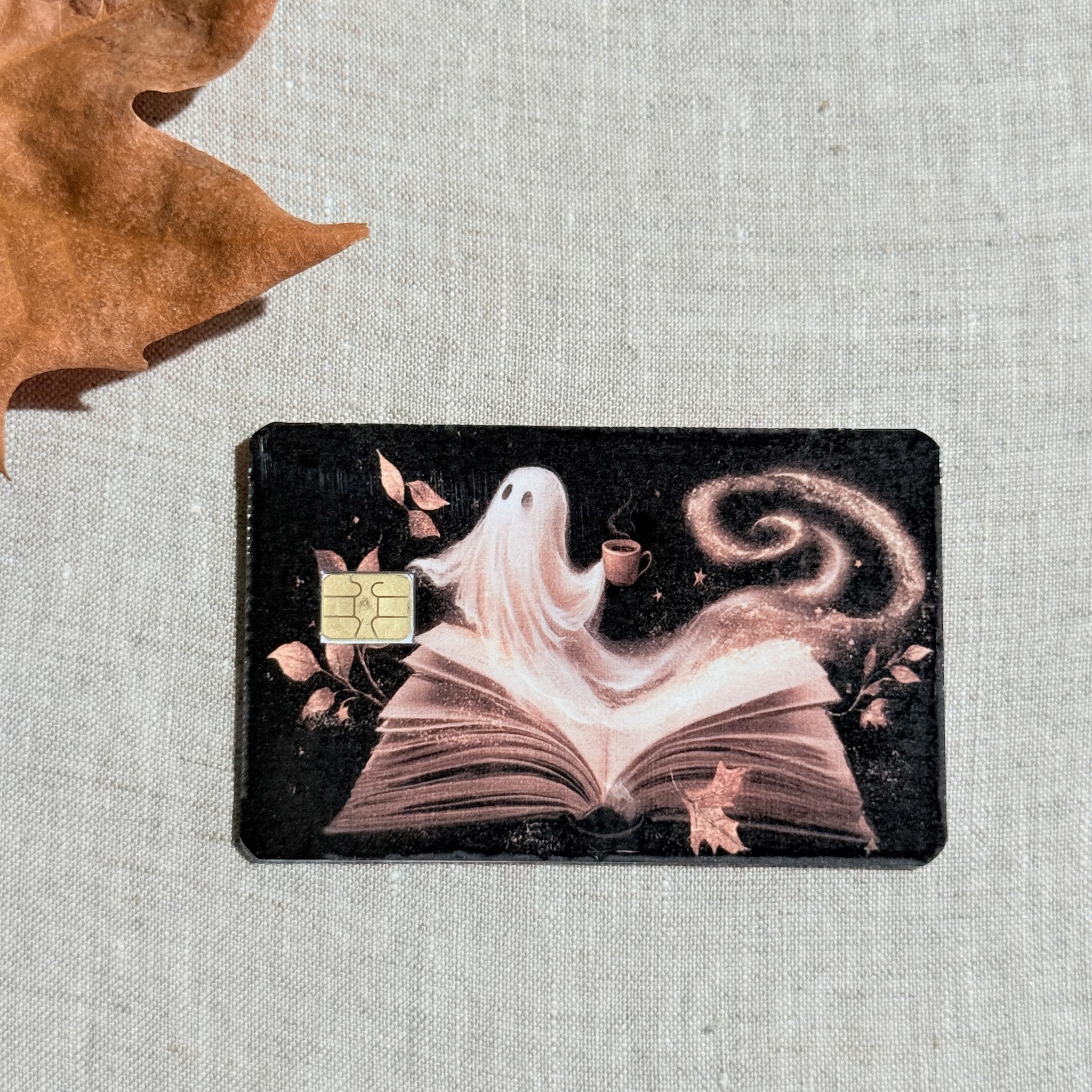 Cozy Halloween ghost debit card sticker shown flat on linen with a fall leaf