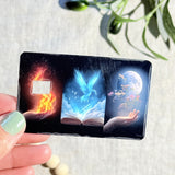 Elemental magic book card skin with fire, blue phoenix and moon panels on a black debit card by The Sticker Era