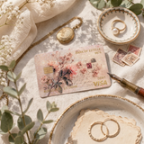 A decorative credit card styled in a flat lay on a cream fabric surface with a gold pocket watch, rings, flowers, stamps, and a fountain pen. The card reads Bank of Estelle and Visa. by the sticker era. 