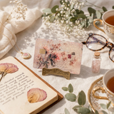 A styled flat-lay featuring a rectangular vintage-style floral credit  card decal on a small gold stand, surrounded by a book with dried flowers, a small vial, glasses, and teacups on a white fabric surface. by the sticker era. 