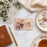A rectangular vintage-style floral debit card skin or credit card  art print with pink roses and script lies on a white marble surface, styled in a flat lay with a brown monogram notebook, gold pen, eucalyptus sprigs, cream fabric, rings, antique keys, and a teacup. by the sticker era