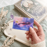 Hand holding celestial violin card skin above cream tray near The Bard's Tale novel BY THE STICKER ERA