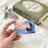 Hand with sage-green manicure and gold rings holding the fantasy card next to a green and gold hardcover novel titled The Whispering Wild and lilac purple blossoms on cream fabric by The Sticker Era