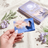 Fingertips with soft lavender polish grip the wolf card near a purple and gilded book called The Moonlit Pack, surrounded by sage sprigs, violet stock florals, and scattered ivory pearls by The Sticker Era