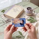 Two hands present the vinyl card above a mint-green wooden table holding a cream and lavender book titled The Moonstone Saga, a purple ceramic mug, dried lavender stems, and fresh garden sage leaves by The Sticker Era