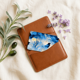 Moody corvid vinyl card cover tucked into the front pocket of a tan leather bifold wallet, arranged on ivory cloth with lavender blooms and sage sprigs BY THE STICKER ERA