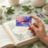 Fingers holding cosmic violin card skin over The Whispering Woods storybook amid sage sprigs BY THE STICKER ERA